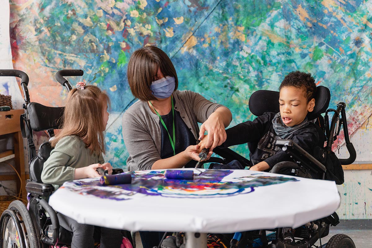 An adult is helping two kids on wheelchair in an art class