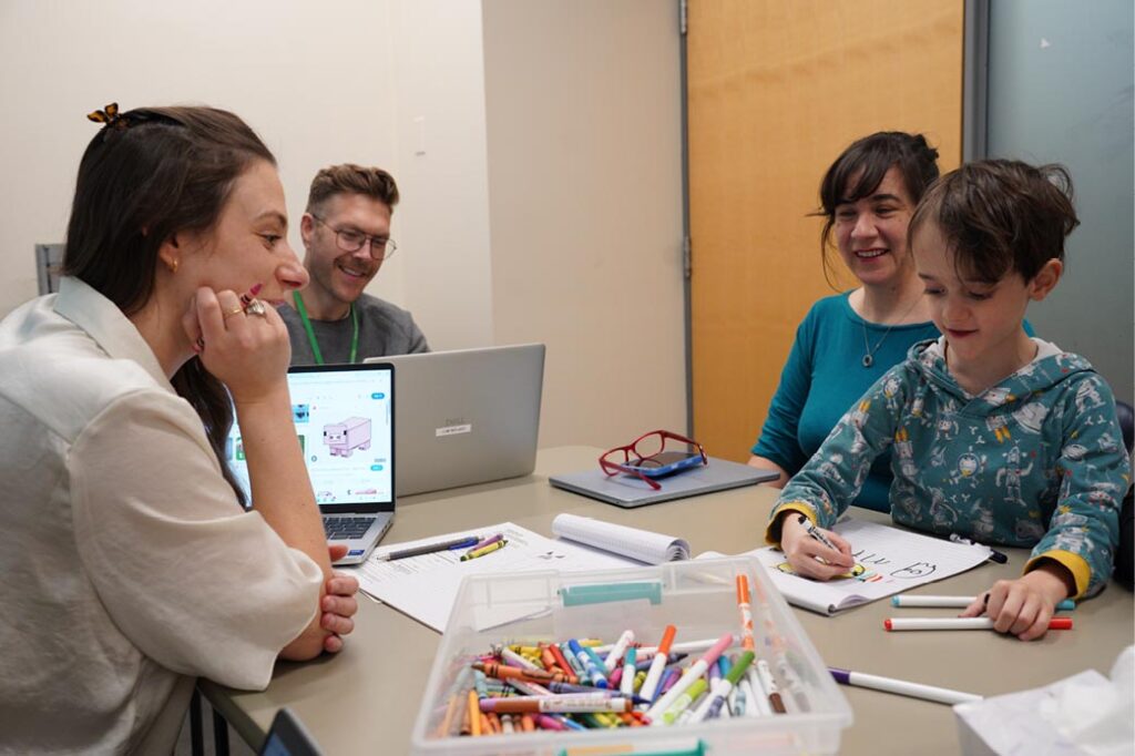 Three adults are accompanying with a kid who is drawing on a paper